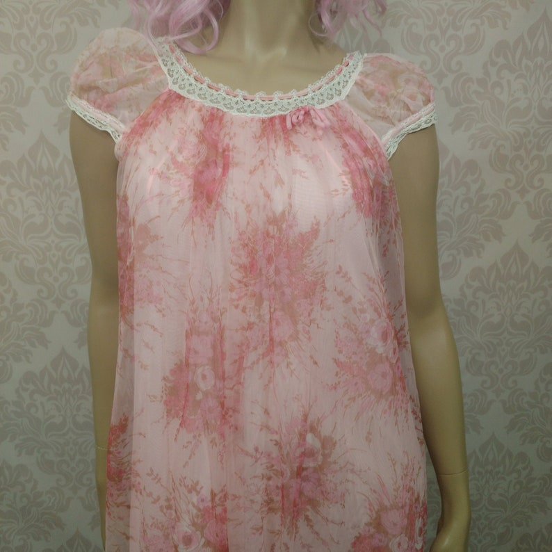 Pode incluir: Uma camisola de noite rosa vintage com um padr&atilde;o floral. O tecido transparente apresenta mangas curtas e uma borda de renda branca ao redor do decote e das mangas. Um pequeno la&ccedil;o est&aacute; no centro do decote.