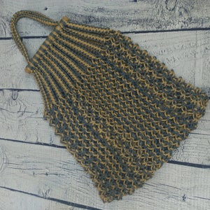 May include: A handmade, open-weave tote bag in a natural beige and blue-gray color scheme. The bag features a looped handle and wooden beads at the top. The bag is made of a woven material.