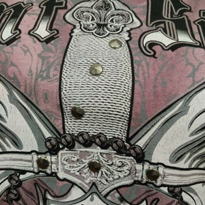 May include: A pink t-shirt with a graphic print of a silver dagger with a fleur-de-lis design. The dagger is surrounded by a silver chain and wings. The shirt has a distressed look.
