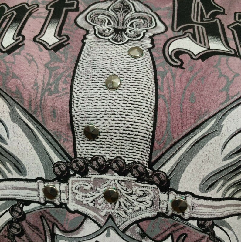 May include: A pink t-shirt with a graphic print of a silver dagger with a fleur-de-lis design. The dagger is surrounded by a silver chain and wings. The shirt has a distressed look.