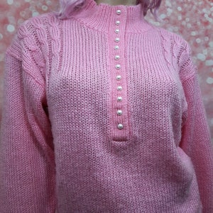 May include: A pink knitted sweater with a mock neck and a row of pearl buttons down the front.