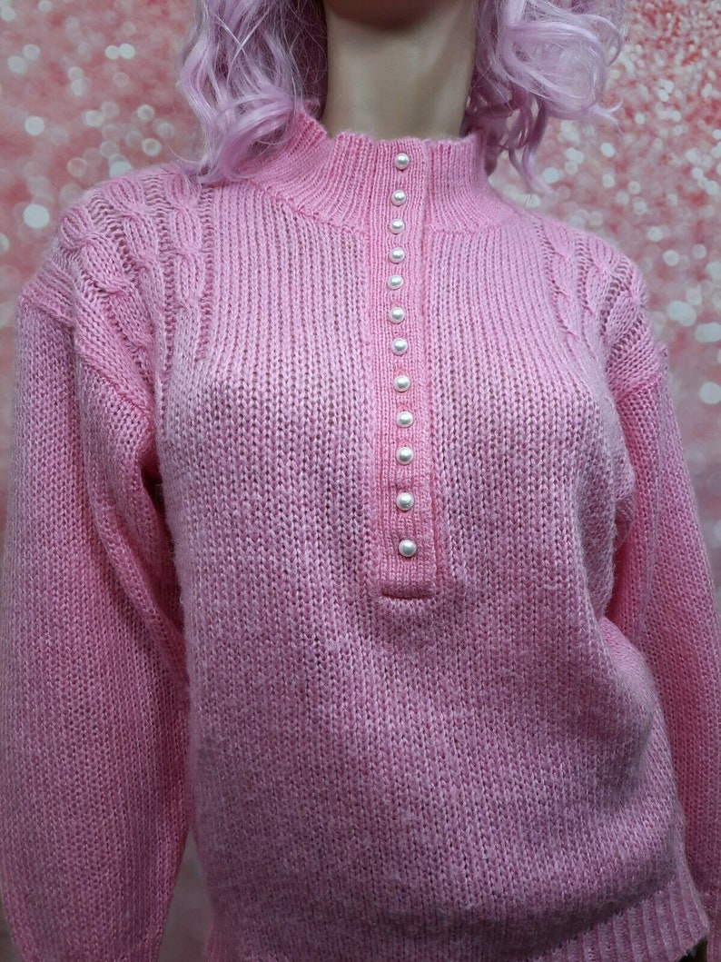 May include: A pink knitted sweater with a mock neck and a row of pearl buttons down the front.