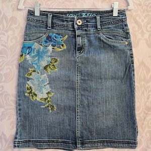 May include: A blue denim skirt with embroidered floral details in shades of blue, green, and white. The skirt has a waistband with the text "Point Zero" in green.
