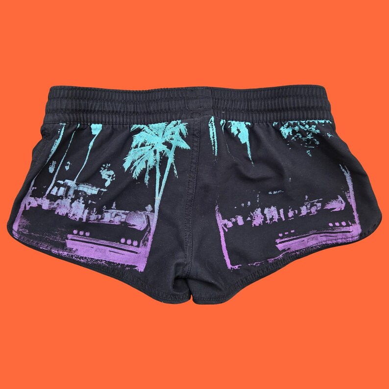 Op de afbeelding: Zwarte sportbroek met een tropisch design. De shorts hebben een paarse en turquoise palmboom- en stadsprint. De tailleband is elastisch. De shorts worden getoond tegen een oranje achtergrond.