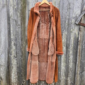 May include: A long brown suede coat with a shearling lining. The coat has a stand-up collar, long sleeves, and a button closure. The coat is open and the lining is visible.