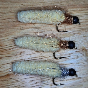 May include: Three handmade fishing flies. Each fly has a cream-colored body, a dark head, and a curved hook. The flies are adorned with gold and silver tinsel and brown thread, resting on a wooden surface.