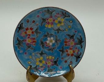 May include: A small round decorative plate featuring a light blue enamel background with a repeating pattern of pink and yellow cherry blossom flowers and dark green leaves, supported by a gold-toned metal display stand with two curved prongs.