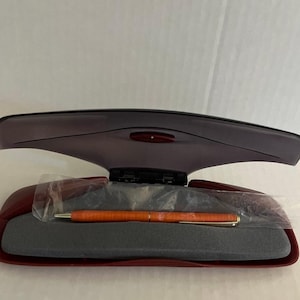 May include: An open, burgundy pen case with a matching lid. Inside, a pen is visible, encased in plastic. The pen is orange with gold accents. The case has a gray, foam-like interior.