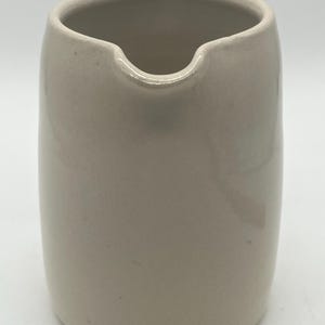 May include: A beige ceramic pitcher with a rounded body and a spout. The pitcher has a smooth, glossy finish and a simple, minimalist design. The opening is wide, and the spout is curved.