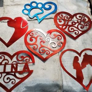 May include: Six metal heart-shaped cutouts, some with intricate designs, others with silhouettes of a dog, a horse, and a person. One heart has the name "Tina" cut out of it.
