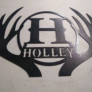 May include: Black metal wall decor featuring a circle with the letter "H" and the name "HOLLEY" inside, surrounded by two stylized antlers.