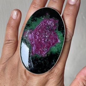 May include: An oval-shaped ring featuring a large, polished zoisite stone with a prominent, textured magenta ruby centre surrounded by deep green and black mineral inclusions, set within a simple, thin silver-toned metal band.