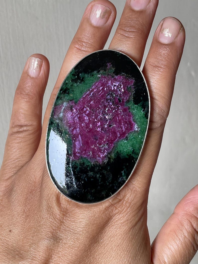 May include: An oval-shaped ring featuring a large, polished zoisite stone with a prominent, textured magenta ruby centre surrounded by deep green and black mineral inclusions, set within a simple, thin silver-toned metal band.