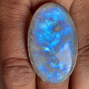 May include: An oval-shaped moonstone ring set in a simple silver band. The gemstone features a translucent white base with a prominent, glowing blue adularescence that shifts across the surface, displaying internal fractures and inclusions.