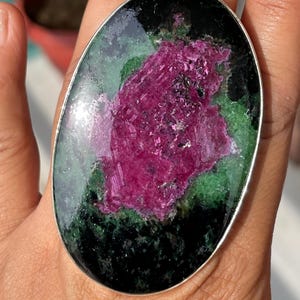 May include: An oval ring featuring a large, polished zoisite stone with a prominent, textured magenta ruby center surrounded by deep green and black mineral inclusions, set within a smooth, thin silver-toned metal band frame.