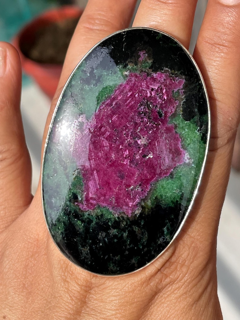May include: An oval ring featuring a large, polished zoisite stone with a prominent, textured magenta ruby centre surrounded by deep green and black mineral inclusions, set within a smooth, thin silver-toned metal band frame.