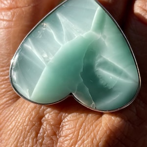 May include: A heart-shaped ring featuring a pale turquoise gemstone with a silver-colored metal band. The gemstone has a unique, textured appearance with white veining. The ring is a piece of jewelry.