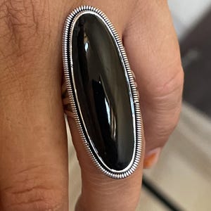 May include: A large, oval-shaped ring with a polished black stone set in a silver-colored metal band. The band has a detailed, textured border. The ring is worn on a finger, showcasing its size and design.