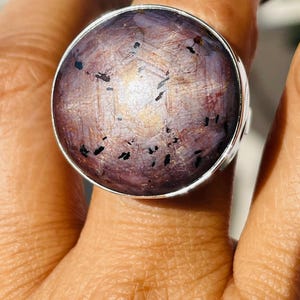 May include: A statement ring with a large, round, purplish-brown gemstone displaying a star effect, set in a silver band. This striking piece of jewellery would add a touch of elegance to any outfit.