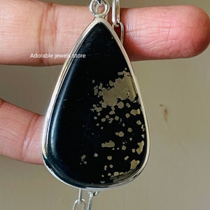May include: A teardrop-shaped pendant with a black stone speckled with gold-colored flecks, set in a silver-colored metal frame. The pendant hangs from a silver chain. The text "Adorable jewelz store" is visible.