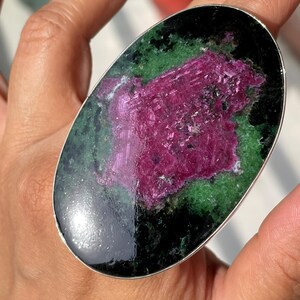 May include: An oval-shaped ring featuring a large, polished gemstone with a vibrant magenta centre surrounded by deep green and black mineral inclusions, set within a smooth, thin silver-toned metal band that encircles the stone's perimeter.