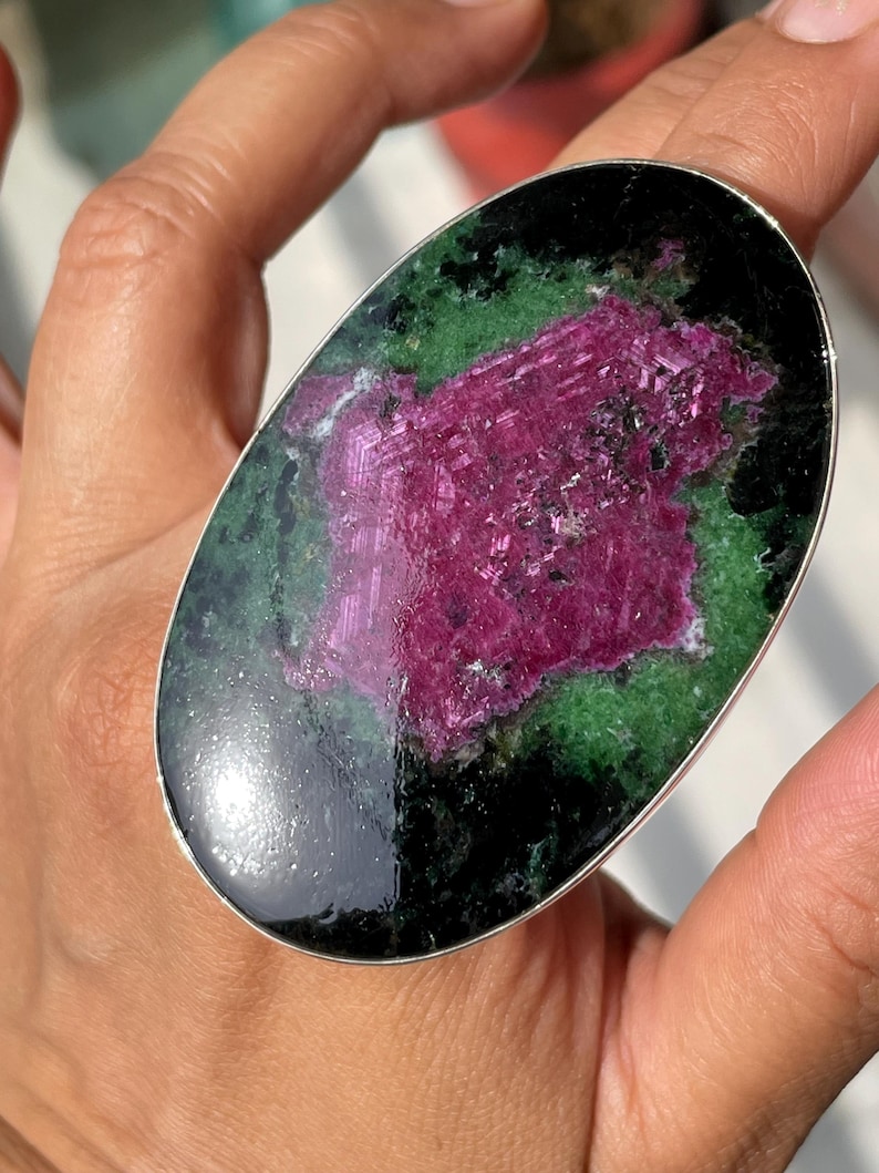 May include: An oval-shaped ring featuring a large, polished gemstone with a vibrant magenta centre surrounded by deep green and black mineral inclusions, set within a smooth, thin silver-toned metal band that encircles the stone's perimeter.