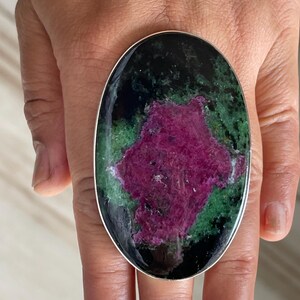 May include: An oval-shaped ring featuring a large, polished gemstone with a vibrant magenta-pink centre surrounded by mottled green and black mineral inclusions, set within a smooth, thin silver-toned metal band.