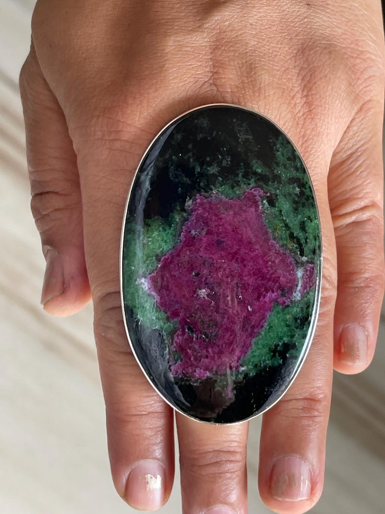 May include: An oval-shaped ring featuring a large, polished gemstone with a vibrant magenta-pink centre surrounded by mottled green and black mineral inclusions, set within a smooth, thin silver-toned metal band.