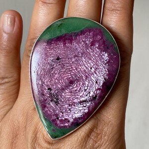 May include: A teardrop-shaped ring featuring a large, polished ruby zoisite gemstone. The stone displays a vibrant magenta-purple centre surrounded by a deep green border, set within a smooth, thin silver-toned metal band that frames the gem.