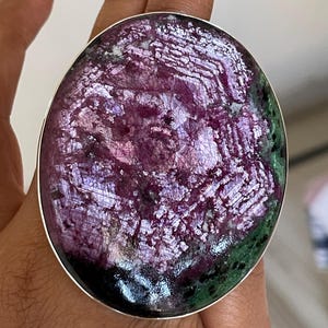 May include: A large, round ring with a polished, iridescent stone. The stone features a mix of deep purple, green, and silver tones, set in a silver band. The ring's design is bold and eye-catching, suitable for statement jewellery.