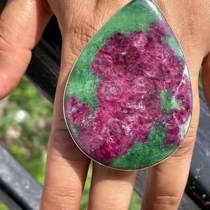 K&ouml;nnte beinhalten: Ein gro&szlig;er tropfenf&ouml;rmiger Ring mit einem polierten Rubin-Zoisit-Edelstein in einer silberfarbenen Metallfassung. Der Stein zeigt eine lebendige Mischung aus tief magentafarbenen Rubinkristallen, die in eine glatte, gr&uuml;ne Zoisit-Matrix eingebettet sind.