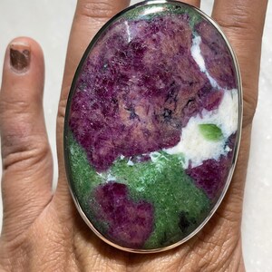 May include: A large oval ring with a polished stone in shades of purple, green, and white, set in a silver-tone metal band. The ring is a bold statement piece, highlighting the natural patterns of the gemstone.