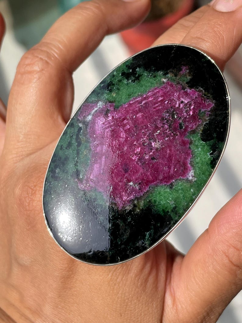 May include: An oval-shaped ring featuring a large, polished gemstone with a vibrant magenta centre surrounded by deep green and black mineral inclusions, set within a smooth, thin silver-toned metal band that encircles the stone's perimeter.