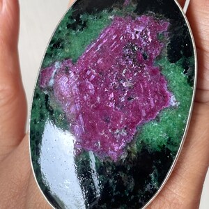 May include: An oval-shaped ring featuring a large, polished ruby zoisite gemstone set in a smooth silver metal bezel. The stone displays a prominent, textured magenta-pink centre surrounded by mottled dark green and black mineral inclusions.