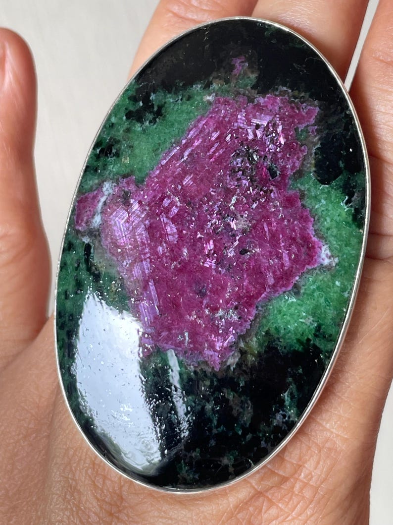 May include: An oval-shaped ring featuring a large, polished ruby zoisite gemstone set in a smooth silver metal bezel. The stone displays a prominent, textured magenta-pink centre surrounded by mottled dark green and black mineral inclusions.