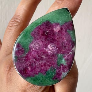 May include: A teardrop-shaped ring featuring a large, polished ruby and zoisite gemstone set in a silver-toned metal frame. The stone displays a vibrant contrast between deep magenta-pink crystalline clusters and a mottled green mineral matrix.