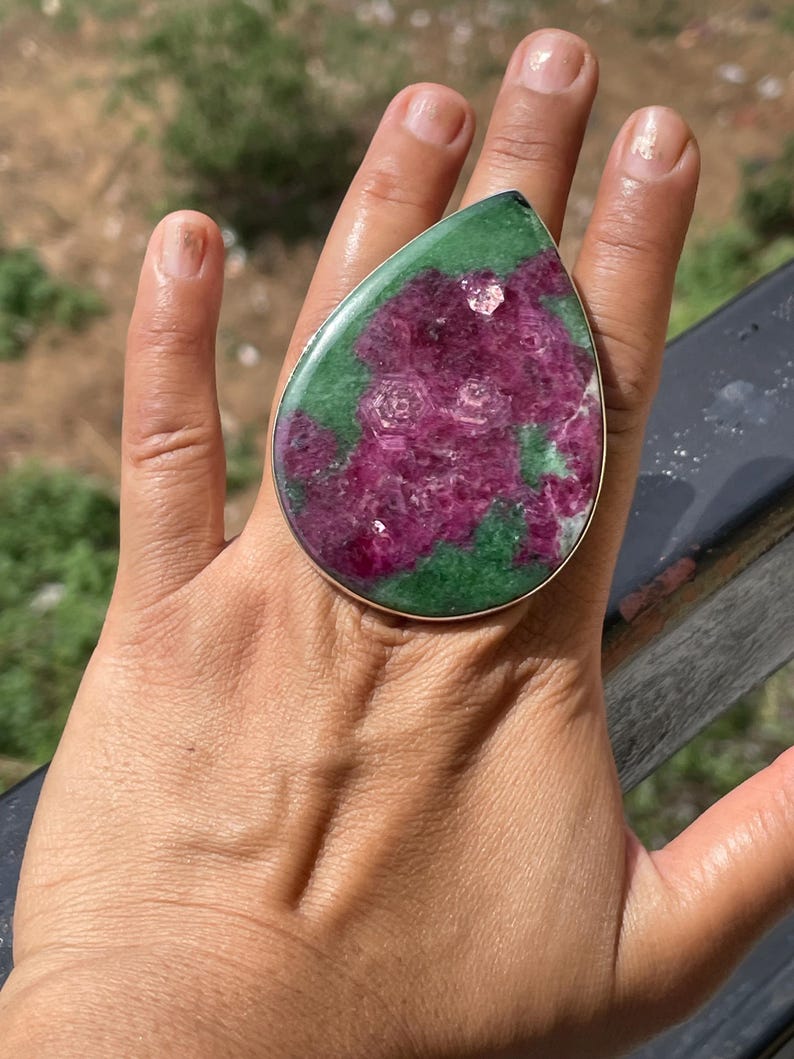 K&ouml;nnte beinhalten: Ein gro&szlig;er tropfenf&ouml;rmiger Ring mit einem polierten Rubin-Zoisit-Edelstein in einer schlichten Fassung aus silberfarbenem Metall. Der Stein zeigt eine lebendige Mischung aus tief magentafarbenen Rubineinschl&uuml;ssen auf einer dunkelgr&uuml;nen Zoisit-Basis.