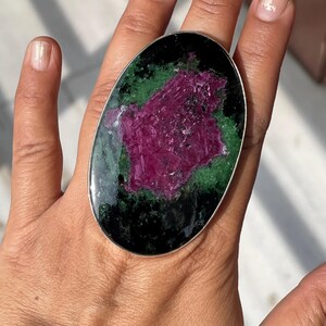 May include: An oval-shaped ring featuring a large, polished zoisite stone with a prominent, textured magenta ruby centre surrounded by deep green and black mineral inclusions, set within a simple, smooth silver-toned metal band frame.