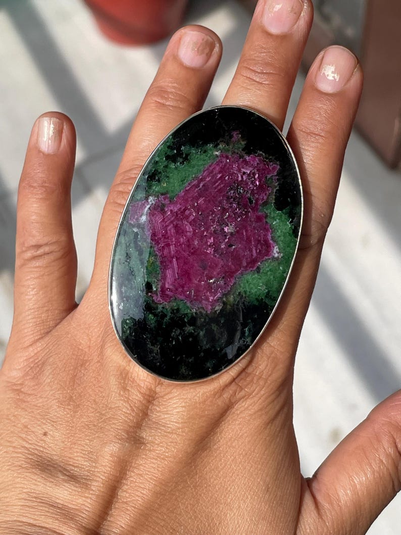 May include: An oval-shaped ring featuring a large, polished zoisite stone with a prominent, textured magenta ruby centre surrounded by deep green and black mineral inclusions, set within a simple, smooth silver-toned metal band frame.