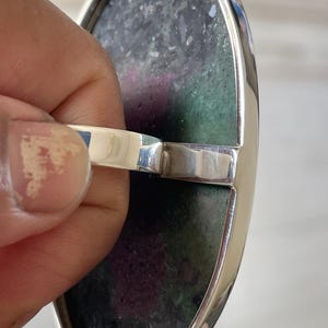 May include: An oval ring featuring a large, flat, polished stone with a mottled texture in shades of deep forest green and vibrant magenta. The stone is set within a smooth, polished silver-toned metal bezel and band, showcasing a clean, reflective finish.