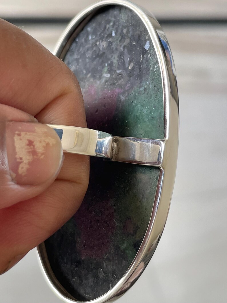 May include: An oval ring featuring a large, flat, polished stone with a mottled texture in shades of deep forest green and vibrant magenta. The stone is set within a smooth, polished silver-toned metal bezel and band, showcasing a clean, reflective finish.
