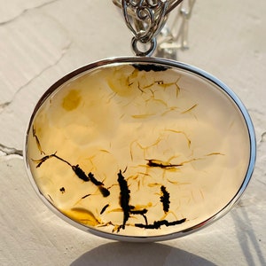 May include: A silver pendant featuring a round, light brown agate stone with black dendritic patterns. The pendant is attached to a silver chain.