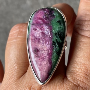 May include: A large, teardrop-shaped ring with a polished ruby zoisite stone. The stone has deep pink and green colours, set in a silver-coloured metal band. The ring is shown on a hand.