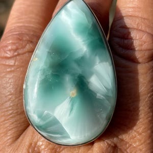 May include: A large, teardrop-shaped ring featuring a polished, light blue gemstone with white veining. The stone is set in a silver-colored metal band. The ring is shown on a hand.