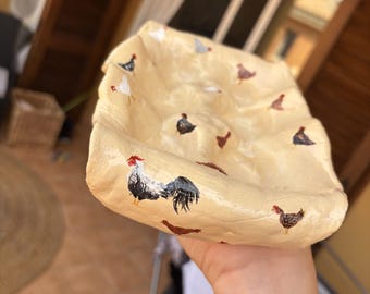 Puede incluir: Un cuenco de cerámica beige pintado a mano con un diseño de gallo y gallina. El cuenco está decorado con varias ilustraciones de pollos en negro, blanco y marrón. El cuenco tiene una forma orgánica única.