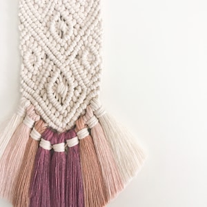 Puede incluir: Un tapiz de macramé blanco con un patrón geométrico y una franja de flecos rosados, rojos y beige.