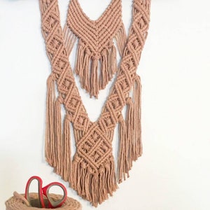 May include: A macrame wall hanging with a V-shape design, featuring a natural wood branch at the top and a light brown color throughout.