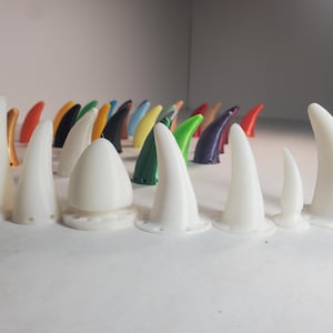 Op de afbeelding: Een verzameling witte, hoornvormige objecten van verschillende grootte met een afgeronde basis. Achter hen bevinden zich kleinere hoorns in verschillende kleuren, waaronder roze, oranje, groen en blauw. De objecten zijn op een wit oppervlak geplaatst.