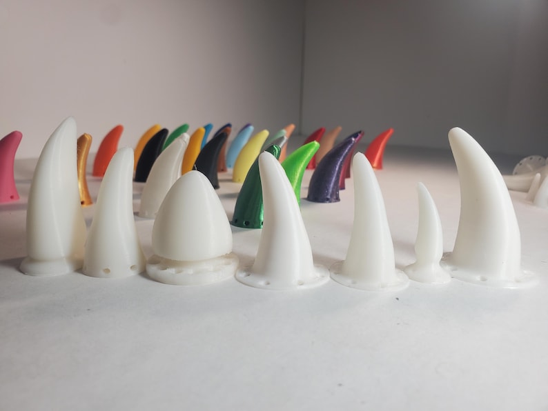 Op de afbeelding: Een verzameling witte, hoornvormige objecten van verschillende grootte met een afgeronde basis. Achter hen bevinden zich kleinere hoorns in verschillende kleuren, waaronder roze, oranje, groen en blauw. De objecten zijn op een wit oppervlak geplaatst.