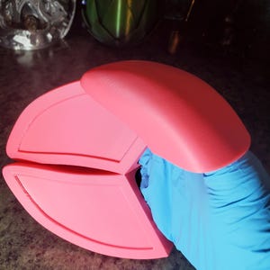 May include: A bright pink, three-part, hinged object, possibly a container or lid, held by a gloved hand. The object has a smooth, matte finish and rounded edges. The background includes other decorative objects.